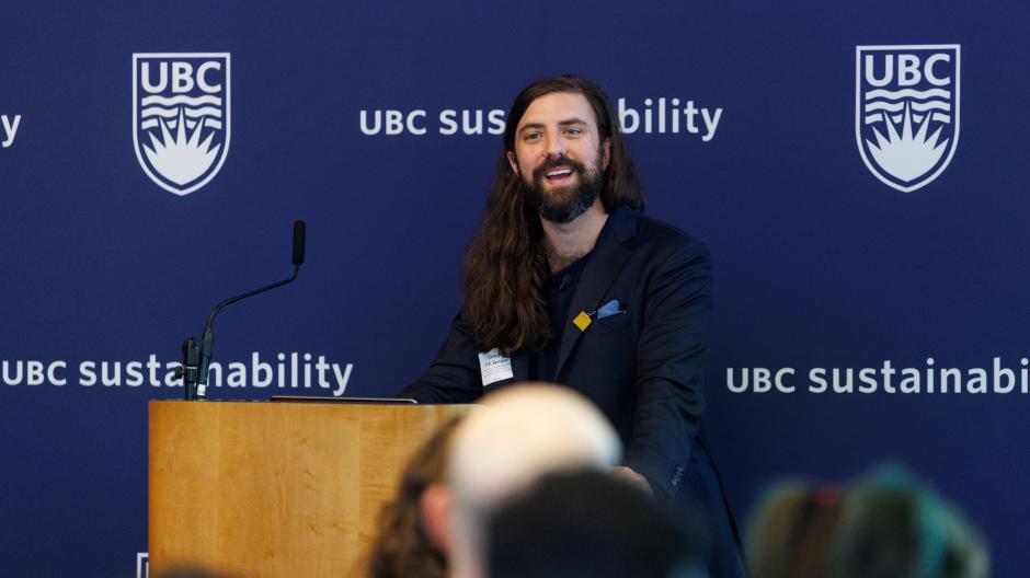 A person with long brown hair and a suit jacket smiling behind a podium and in front of a blue backdrop.
