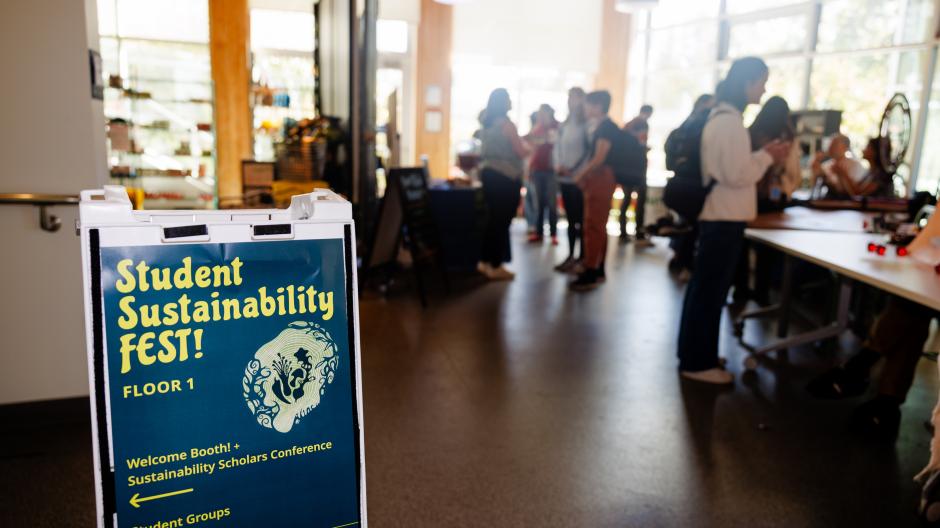 A poster on a stand with the title 'Student Sustainability FEST!' in the foreground with a blurred scattering of people in the background.