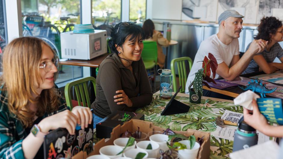 Three people seated behind tables with informative displays smiling to and talking with people standing on the other side of the tables.