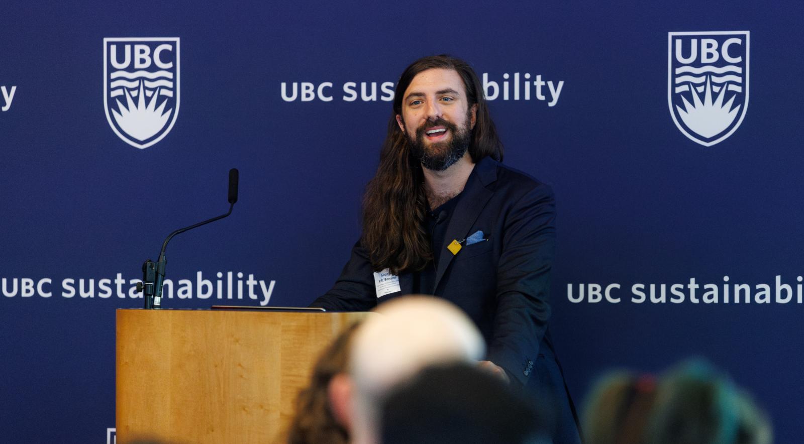 A person with long brown hair and a suit jacket smiling behind a podium and in front of a blue backdrop.