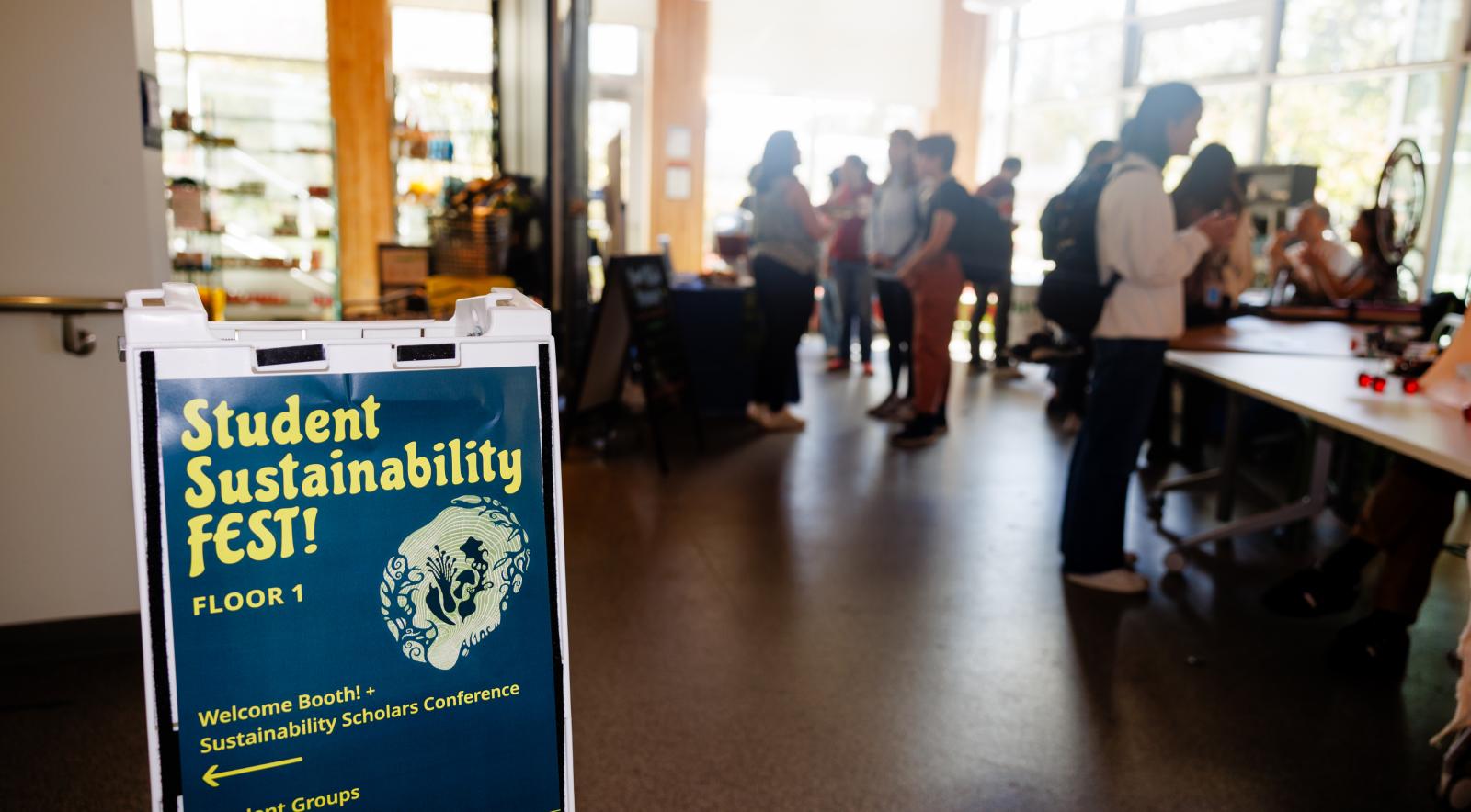 A poster on a stand with the title 'Student Sustainability FEST!' in the foreground with a blurred scattering of people in the background.