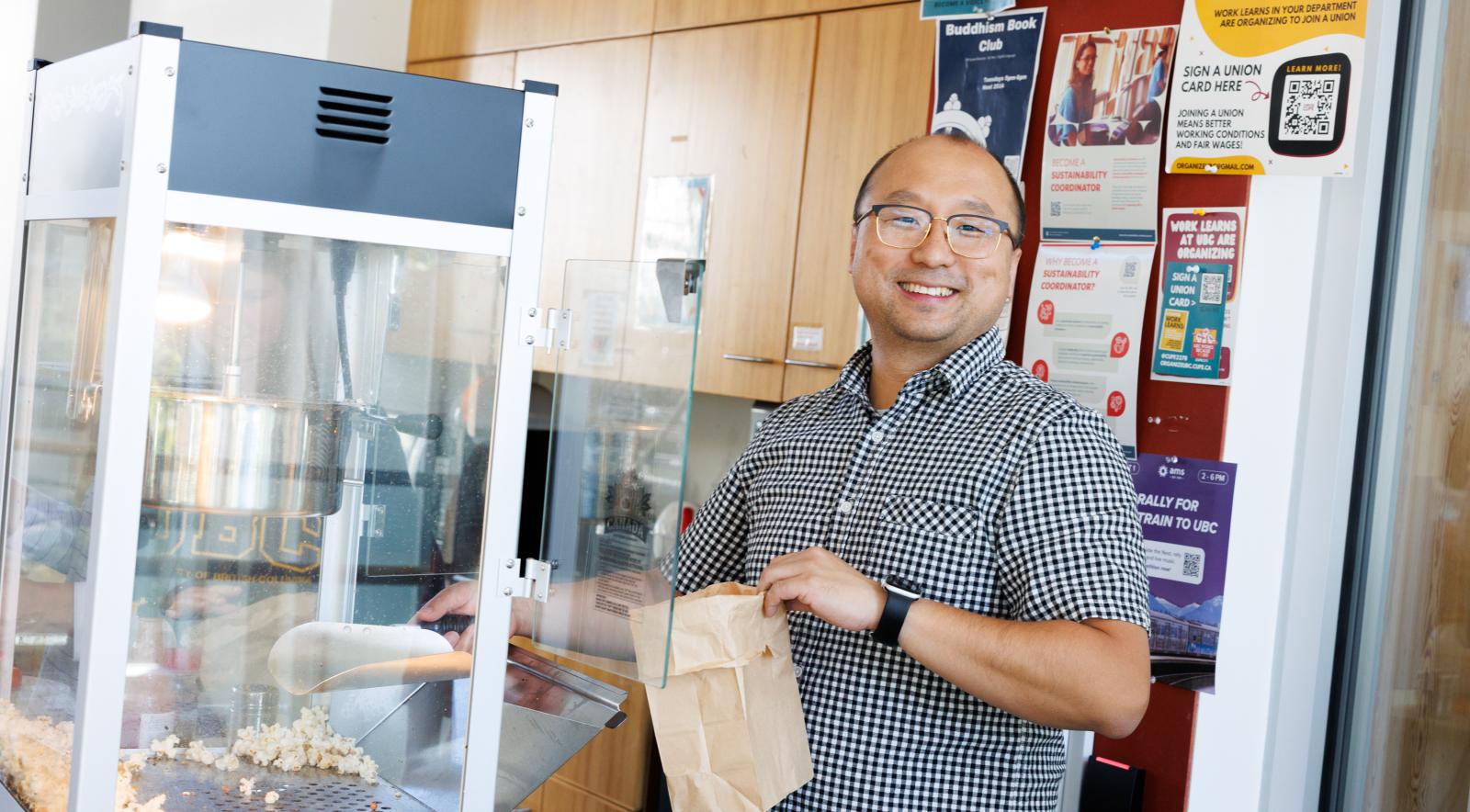 A person smiling and scooping fresh popcorn from a popcorn machine into a brown paper bag.