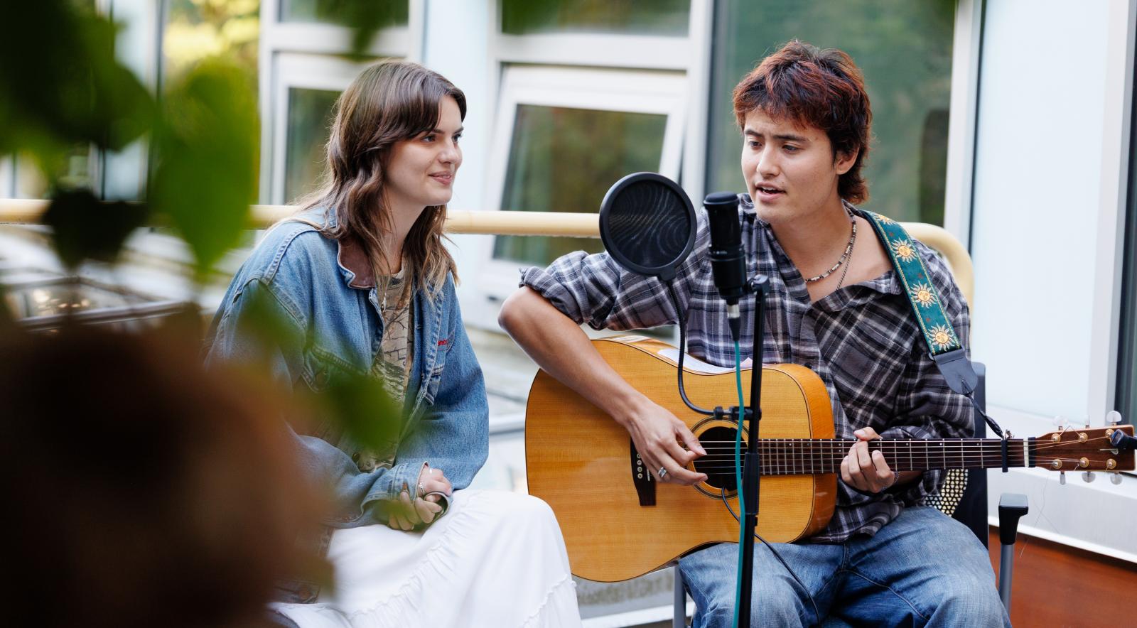 Two people sitting by a microphone. One is signing and the other is playing the guitar and singing.